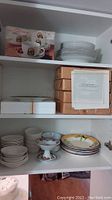 Shelf showing cappuccino cups and saucers in boxes, Royal Doulton plates stacked, boxed collector plates, and assorted plates and bowls.