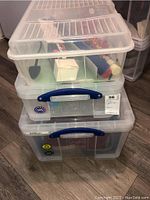 Three stacked clear plastic storage bins with blue handles, containing various household items