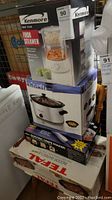 Photo showing boxed Kenmore two tier food steamer on top, Brevetti crock pot below, Tfal indoor BBQ box at bottom, and Salton crepe maker box in stack.