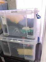 Two stacked translucent plastic bins filled with assorted folded ladies clothing, tagged with labels showing 'Summer Dresses & Skirts' and 'New with or without tags'.