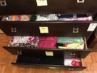 Three open drawers of dark wood dresser filled with folded women's tops in various colors and prints.