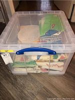 Clear plastic storage bin with blue handle filled with various folded kitchen linen items including doilies and towels.