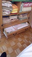 Wooden shelving with three levels holding folded linens including face cloths, tablecloths, and blankets, with a drawer below containing additional linens.