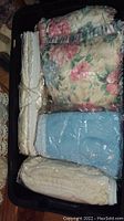 Top view of black plastic bin filled with folded blankets and sheets, including floral patterned blankets, blue plastic wrapped blanket, and tied cream sheets.