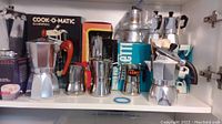 Shelf with multiple aluminum stovetop coffee percolators and espresso makers, some in boxes, showing variation in design and size.