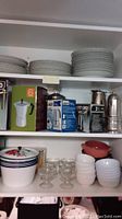 Shelves of white plates, boxed Bialetti espresso makers, glass dessert bowls, small ceramic bowls, and decorative casserole pot with floral pattern.