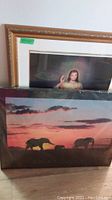 Partial view of two framed items stacked: one print showing a young child, the other a poster with elephants at sunset.