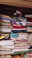 Shelf filled with stacked folded fabric tablecloths, napkins, and linens in a range of colors including pastels, reds, and neutrals.