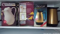 Photograph of three beverage dispensers: two boxed Connoisseurs insulated beverage servers and one Hot'n Cold dispenser unboxed, showing appearance and packaging.