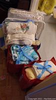 Stack of folded towels, linens, bedspread, and sheets with some items packaged in red bags and others folded on top of a suitcase.