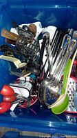 Top view of blue bin containing assorted kitchen utensils including metal spoons, wooden spoons, tongs, and graters.