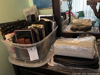 Bin filled with assorted wallets, evening clutches, and makeup bags, some wrapped in plastic, stacked on a dark surface showing quantity and variety