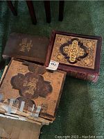 Three antique bibles grouped on green carpet: one large heavily embossed bible with gold floral details, one large brown bible with gold text, one bible with damaged cover taped.