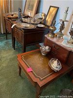 Three antique wooden tables shown together with various silver items on top. Two end tables with curved fronts and two drawers each. Larger table with pink marble top and leather inlay writing surface pulled out.