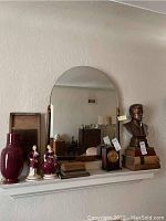 Long shot of mantel shelf displaying the Art Deco mirror with arched top and carved wood sides. Various decorative items also present.