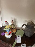 Photo showing two clown music boxes, ornate metal coffee pot, and green handheld mirror on wooden surface.
