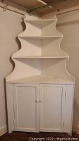 Corner cabinet in a room corner, showing the closed cabinet doors and three open shelves above, painted white with signs of wear and scratches.