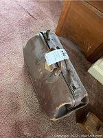 Exterior view of the vintage leather doctor's bag resting on carpeted floor, showing wear and patina on leather surface and handle with a tag attached.
