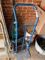 Blue steel dolly standing upright against a brick wall with a lot sticker on handle.