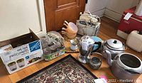 Wide view of all kitchen items including kettles, glass dishes, box of canning jars, and hand mixer on floor.