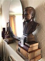 Photo of bronze-tone bust of John F. Kennedy on vintage books placed on mantle with mirror reflecting background