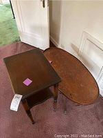Two wooden tables placed side by side: one oval coffee table with tripod base and claw feet, and one rectangular small lamp table with lower shelf; visible wear on surfaces.