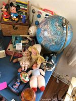 Photo showing assorted vintage kids toys and items including dolls, a globe, and Mickey and Minnie Mouse figurines on a blue table.