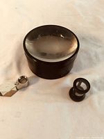 Three vintage magnifying loupes displayed on a white surface including a large round black magnifier, small black loupe, and silver folding loupe.