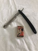 Full view of the antique men's straight razor with black handle alongside a box of vintage Don Juan Blades on a white cloth background.