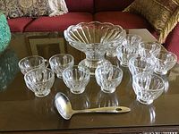 Clear glass fruit bowl, 12 matching glass cups, and a metal ladle arranged on a glass table.