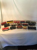 Full view of 15 model trains laid out on a white cloth background showing various types and colors including locomotives, passenger and freight cars.