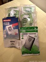 Photo of multiple vacuum bag packages arranged on a surface, showing Miele and Crucial brands, plus one Kenmore/Panasonic package.