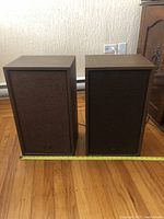 Front view of pair Akai speakers side by side on wooden floor, showing fabric grille and Akai logo.