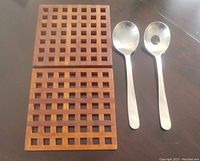 Two teak trivets with open grid design placed next to a pair of stainless steel salad servers on a dark reflective surface