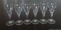 Six clear Georgian-style stemmed sherry or port glasses lined in a row on dark surface, clean with no visible damage.