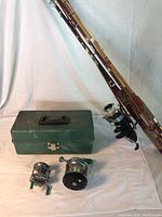 Fishing rods leaning against backdrop next to green tackle box and two fishing reels