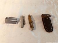 Photo showing all four items: lighter, two knives, and leather sheath laid out on a surface.