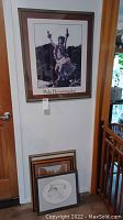 Photo showing four framed prints: one Native Hawaiian dancer print hung on the wall above three other framed prints resting on the floor including a large framed black and white drawing of an owl, a horse drawing, and an Arizona print.