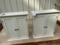 Two white wooden bathroom cabinets side by side, showing front and top angles.