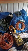 Mixed assortment of extension cords in orange, black, yellow and gray hues laid out on blue tarp, displaying coils and plugs along with a Yellow Jacket power strip in the center.