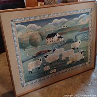 Full view of the framed barnyard print showing sheep, farmhouse, hills, and decorative border mat inside the light wooden frame.