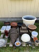 All items in lot displayed on wood pallets outside against a yellow wall, showing full variety of plant pots in ceramic, plastic, wicker along with other decorative gardening items.