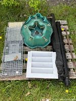 Items arranged on a wooden pallet including live trap cage, vent cover, rolled fencing, tree stand bases, wooden sticks and hardware box.