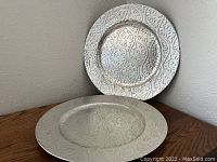 Two white ceramic plates with an embossed floral and vine pattern, stacked with one leaning on the wall