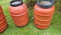 Two orange plastic water barrels shown side-by-side on grass. Both have black screw-on lids and visible valves and hose fittings near base.
