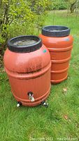 Full view of two orange water barrels outside on grass, showing large black screw lids and orange ribbed bodies.