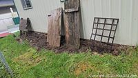 Photo showing weathered wooden barn door planks leaning against a wall with a metal grid piece on the ground.