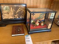 Photo of the two glass cases and the wooden-covered book on a wood surface, showing the carved village and duck figurines