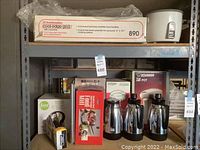 Wide view of entire lot stacked on metal shelving including boxed Toastmaster cool-edge grill on top shelf, Zojirushi rice cooker, Food Network kettle, Zojirushi airpot, KitchenAid mixer box and trash bags on lower shelf along with three chrome pitchers or airpots.