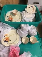 General overview showing a large teal bin filled with octagonal plates, cups, and wrapped items with floral patterns.